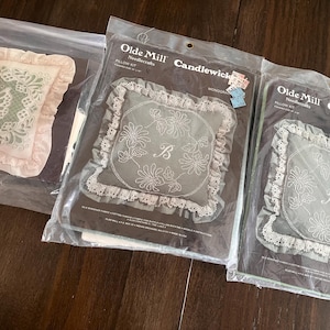 May include: Three pillow kits in plastic packaging. The kits feature embroidered designs, including a butterfly and floral patterns. The pillows have ruffled edges and are labeled "Olde Mill Candlewick Needlecrafts".