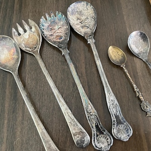 May include: A collection of six silver-toned metal utensils including two large serving spoons, one salad fork, one small decorative spoon with a twisted handle, and two smaller spoons. Each piece features tarnished surfaces and ornate detailing.