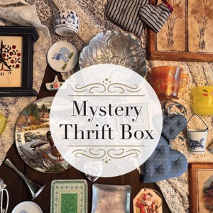 May include: A collection of vintage items, including a clock, plates, a decorative dish, and mugs. The image features a variety of colors and textures, with the text "Mystery Thrift Box" in the center. The items are arranged on a lace tablecloth.