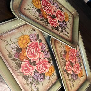 May include: A set of three rectangular serving trays with a light green border and a central floral illustration featuring pink roses, orange daisies, and small purple flowers. The trays have a smooth finish and a slightly raised edge for containment.