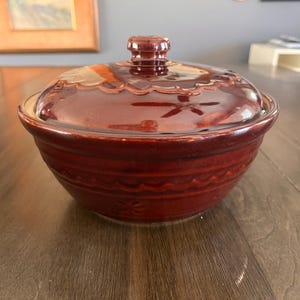 May include: A covered, maroon-colored ceramic bowl with a matching lid. The lid has a decorative scalloped edge and a small knob. The bowl has a floral design and horizontal ridges. A kitchenware item.