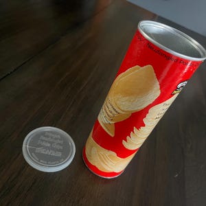 May include: A red cylindrical Pringles Newfangled Potato Chips container with a graphic of stacked chips on the side and a separate clear plastic lid featuring the text Pringles Newfangled Potato Chips printed in white on the top surface.
