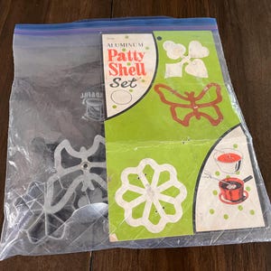 May include: A vintage aluminum Patty Shell Set in a clear plastic bag. The packaging features a green background with illustrations of patty shells and a butterfly shape. The set includes various cookie cutter shapes.
