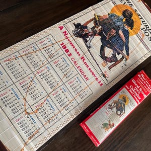 May include: A vintage 1983 calendar featuring a Norman Rockwell illustration, with a bamboo-style backing. The calendar displays the months and days, and is accompanied by its original red box. The box reads "Wood Scroll Calendar and Wall Hanging."