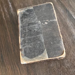 May include: A small, rectangular, vintage book with a dark cover, held together with clear tape. The edges of the book are worn and show signs of age. The book is resting on a wooden surface.