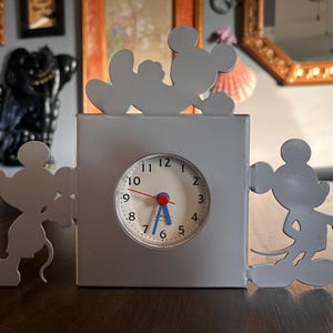 May include: A gray Mickey Mouse-themed clock with a white clock face, red second hand, and blue hour and minute hands. The clock features Mickey Mouse silhouettes on the sides and top. The clock is on a wooden surface.