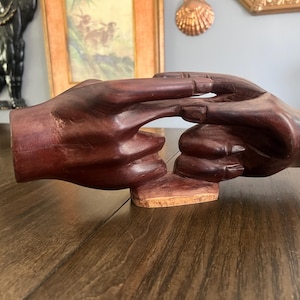 May include: A dark brown wooden sculpture depicting two hands joined together in a clasping gesture. The hands are carved from a single piece of wood with a smooth, polished finish, resting on a small, light-colored, natural-edged wooden base.