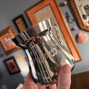 May include: A polished silver cup with a handle and a flared rim. The cup has an engraved design on the front. The cup is held in a hand, with a blurred background of framed artwork and decor.