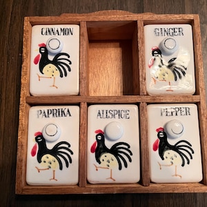 May include: A wooden spice rack with six white ceramic spice jars. Each jar is labeled with a spice name and features a rooster illustration. The spices are cinnamon, ginger, paprika, allspice, and pepper.