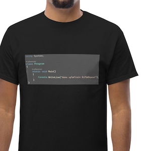 Könnte beinhalten: Ein schwarzes T-Shirt mit einem Code-Snippet, das auf der Vorderseite aufgedruckt ist. Der Code-Snippet befindet sich in einem dunkelgrauen Kasten mit hellgrauem Hintergrund. Der Code-Snippet lautet: "public static void main(String[] args) {System.out.println("Hello World");}"
