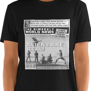 Puede incluir: Camiseta gráfica en blanco y negro con un titular de periódico que dice "ALIENS ARE REAL!" con un titular más pequeño que dice "PRESIDENT IS A TIME TRAVELING CYBORG!" El periódico también presenta un titular más pequeño que dice "10 WAYS TO KNOW IF YOUR PET IS A DEMON".