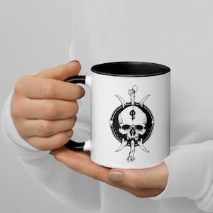 Peut inclure: Une tasse en céramique blanche avec un bord et une poignée noirs. La tasse présente un motif noir et blanc d'un crâne avec un symbole sur son front, entouré d'os.