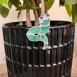 May include: A green and silver stained glass frog plant stake. The frog has large eyes and is attached to a metal stake. The frog is placed in a black ceramic planter with a ribbed design, with a green plant in the background.