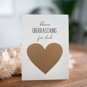 Pu&ograve; includere: Un biglietto bianco con il testo "kleine &Uuml;BERRASCHUNG f&uuml;r dich" e una forma di cuore dorata.