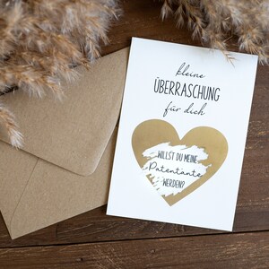 Pu&ograve; includere: Un biglietto bianco con un cuore dorato che dice "kleine &Uuml;BERRASCHUNG f&uuml;r dich" e "WILLST DU MEINE PATENTANTE WERDEN?" all'interno del cuore.