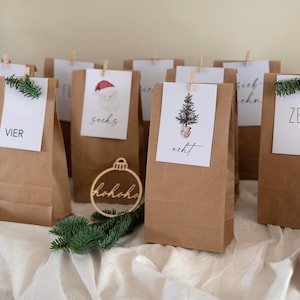 May include: A collection of brown paper bags, each with a white tag clipped on, forming an advent calendar. Some tags have numbers, a Santa illustration, or a Christmas tree. Greenery and a wooden ornament add to the festive theme.