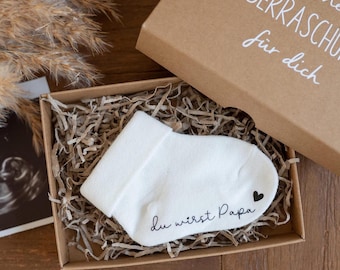May include: A small white sock with the text "du wirst Papa" and a small heart printed on it. The sock is in a brown box with shredded paper. The box has the text "kleine Überraschung für dich" on the top.
