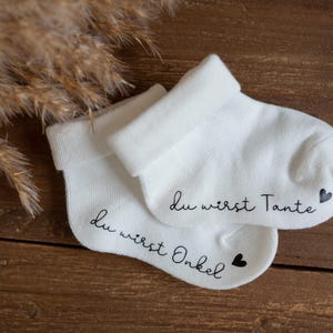 May include: Two white baby socks with black text. One sock says "Du wirst Onkel" with a small black heart. The other sock says "Du wirst Tante" with a small black heart.