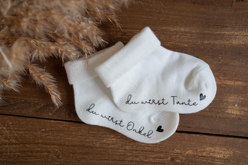 May include: Two white baby socks with black text. One sock says "Du wirst Onkel" with a small black heart. The other sock says "Du wirst Tante" with a small black heart.