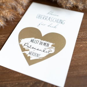 Pu&ograve; includere: Un biglietto bianco con una forma di cuore dorata. Il biglietto dice "Kleine &Uuml;berraschung f&uuml;r dich" e "Willst du mein Patenonkel werden?"
