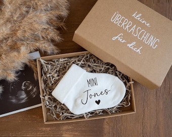 Könnte beinhalten: Ein weißer Babysocken mit dem Namen "Mini Jones" und einem Herz, präsentiert in einer Pappschachtel mit dem Text "kleine Überraschung für dich". Die Schachtel ist mit Papier gefüllt und liegt neben einem Ultraschallbild.
