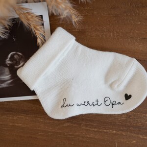 May include: White baby sock with the text "Du wirst Opa" and a small black heart on the heel.