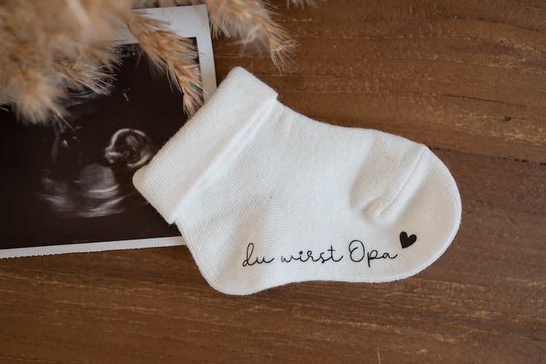 May include: White baby sock with the text "Du wirst Opa" and a small black heart on the heel.