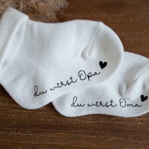 May include: Two white socks with black text and a small black heart. The text on one sock reads "Du wirst Opa" and the text on the other sock reads "Du wirst Oma".