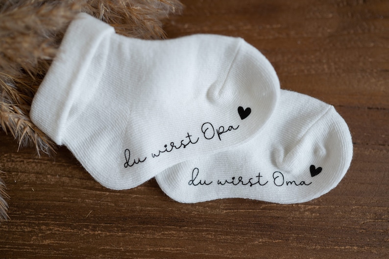 May include: Two white socks with black text and a small black heart. The text on one sock reads "Du wirst Opa" and the text on the other sock reads "Du wirst Oma".