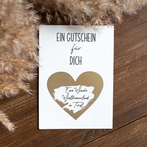 May include: A white card with the text "EIN GUTSCHEIN f&uuml;r DICH" and a gold heart with the text "Eine Woche Wellnessurlaub in Tirol" written on it. The card is on a brown wooden surface.