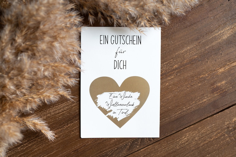 May include: A white card with the text "EIN GUTSCHEIN f&uuml;r DICH" and a gold heart with the text "Eine Woche Wellnessurlaub in Tirol" written on it. The card is on a brown wooden surface.