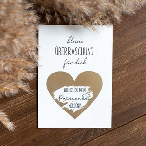 Pu&ograve; includere: Un biglietto bianco con il testo "kleine &Uuml;BERRASCHUNG f&uuml;r dich" e un cuore dorato con il testo "WILLST DU MEIN PATENONKEL WERDEN?" su una superficie di legno marrone.