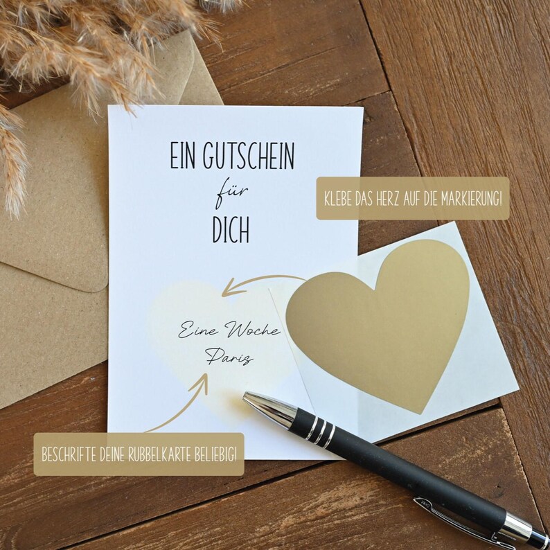 May include: A white card with the text "EIN GUTSCHEIN f&uuml;r DICH" and "Eine Woche Paris" written on it. A gold heart sticker is placed on the card with instructions to "KLEBE DAS HERZ AUF DIE MARKIERUNG!" and "BESCHRIFTE DEINE RUBBELKARTE BELIEBIGI!"