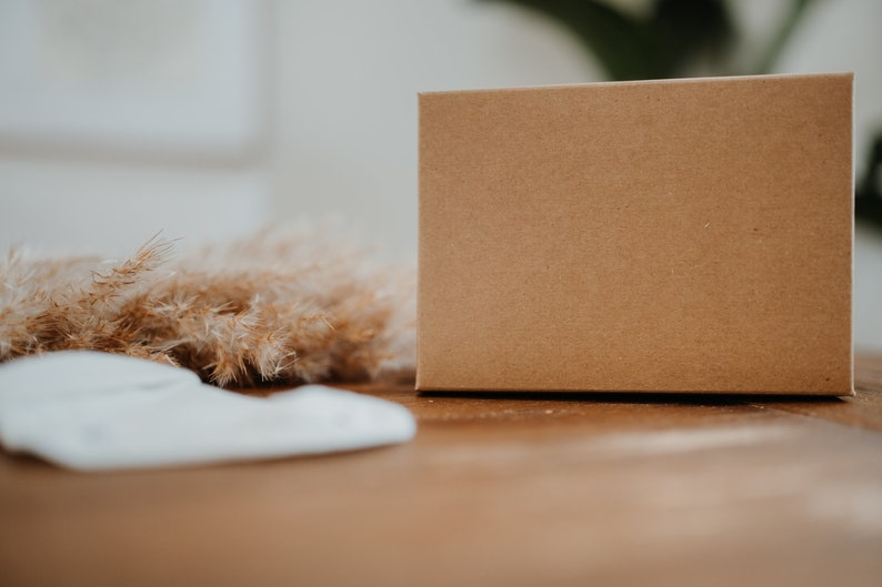 May include: A brown cardboard box sits on a wooden surface with dried grass in the foreground. The box is plain and has no markings.