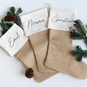 Könnte beinhalten: Drei beige Weihnachtsstrümpfe mit weißen Oberteilen, jeweils personalisiert mit einem Namen in schwarzer Schrift: "Emil", "Mama" und "Christina". Die Strümpfe sind mit Tannennadeln und Tannenzapfen verziert.