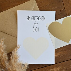 May include: A white card with black text that reads "EIN GUTSCHEIN f&uuml;r DICH". A gold heart sticker is on the right side of the card. A brown envelope is behind the card.