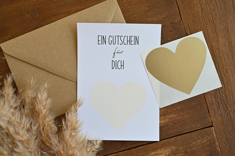 May include: A white card with black text that reads "EIN GUTSCHEIN f&uuml;r DICH". A gold heart sticker is on the right side of the card. A brown envelope is behind the card.