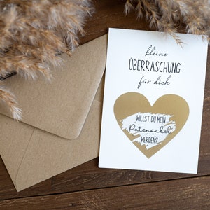 Pu&ograve; includere: Un biglietto bianco con un cuore dorato e il testo "kleine &Uuml;BERRASCHUNG f&uuml;r dich" e "WILLST DU MEIN PATENONKEL WERDEN?"