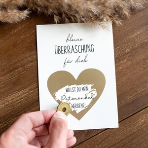 Pu&ograve; includere: Un biglietto bianco con il testo "kleine &Uuml;BERRASCHUNG f&uuml;r dich" e un cuore dorato con il testo "WILLST DU MEIN PATENONKEL WERDEN?" Una moneta d'oro viene utilizzata per grattare il cuore dorato e rivelare il testo.