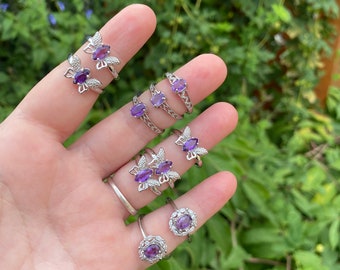May include: A set of six silver rings with purple amethyst stones. The rings feature different designs, including butterfly motifs and a ring with a halo of smaller stones.