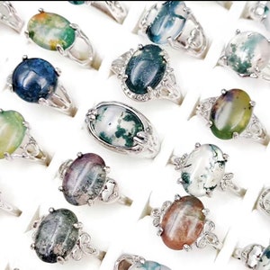 May include: A collection of silver rings with oval-shaped agate gemstones. The stones are a variety of colors, including green, brown, white, and black.