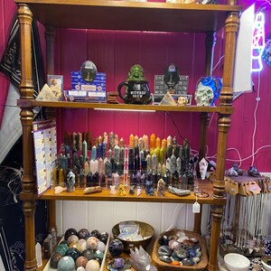 May include: A wooden shelf unit with various crystals, stones, and other magical items. The shelf is decorated with a green witch figurine holding a mug that says "Witches Brew".