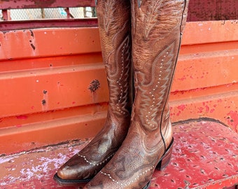 May include: A pair of brown leather cowboy boots with intricate stitching and a pointed toe. The boots have a tall shaft and a stacked heel, showcasing a classic Western design. The boots are set against a red background.