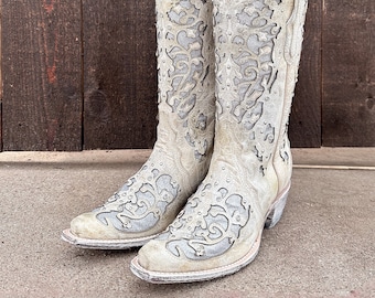 Könnte beinhalten: Ein Paar weiße Cowboy-Stiefel mit aufwendigen floralen Ausschnitten und silbernen Verzierungen. Die Stiefel haben eine spitze Zehenpartie und einen niedrigen Absatz.