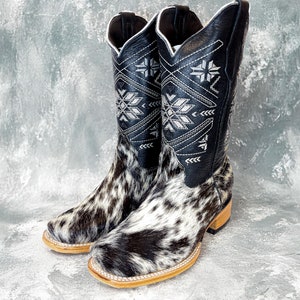 May include: A pair of black cowboy boots with white and black cowhide pattern. The boots have a geometric design embroidered on the shaft in white thread.