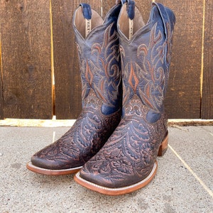 May include: A pair of brown leather cowboy boots with intricate floral embroidery in shades of blue and brown. The boots have a pointed toe and a low heel.