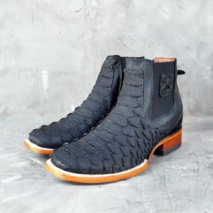 May include: A pair of black leather ankle boots with a unique scale-like texture. The boots have a square toe, a brown and orange sole, and a small decorative emblem on the side. The boots are approximately 6 inches tall.