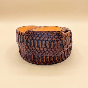 May include: A brown leather belt with a snakeskin pattern. The belt has a buckle and a light brown interior. The snakeskin pattern is dark brown and black. The belt is a fashion accessory.