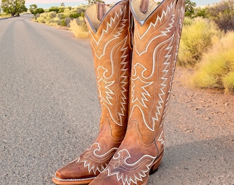 May include: A pair of brown leather cowboy boots with white stitching and a pointed toe. The boots have a decorative eagle design on the shaft.