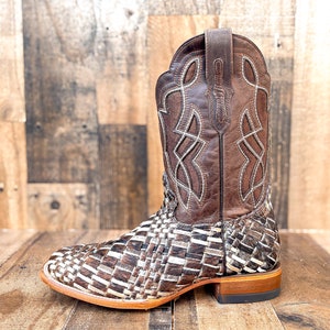 Puede incluir: Una bota de vaquero de cuero marr&oacute;n con un dise&ntilde;o tejido en el pie y una parte superior de cuero marr&oacute;n. La bota tiene costuras decorativas y una puntera cuadrada. El tac&oacute;n mide aproximadamente 3,8 cm de alto.